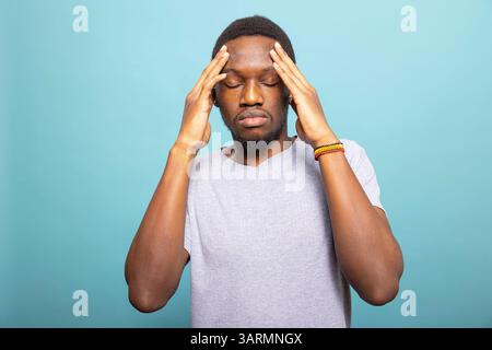 Exhausted black male model with eyes closed suffering from a migraine, rubbing his temples in distress. African american man feeling unwell and overwhelmed by pain, standing against isolated backdrop. Stock Photo