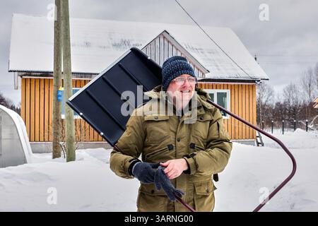 During routine outdoor task involving snow removal older adult male in his 50s looks serious holding shovel beside rural home surrounded by deep snow. Stock Photo