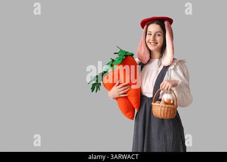 Stylish woman with bunny ears smiling and holding carrot toy and basket with Easter bread on grey background. Easter celebration Stock Photo