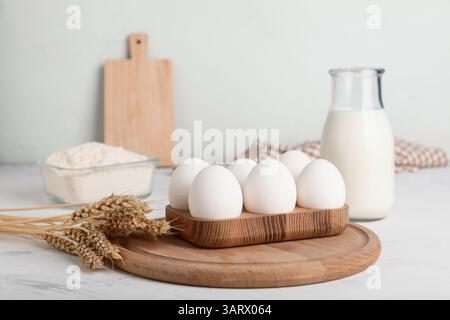 Wooden holder with fresh chicken eggs, bottle of milk, flour and wheat spikelets on light table Stock Photo