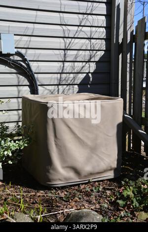 An outdoor central air conditioning condenser unit is covered with a protective beige storage cover outside a residential house. Stock Photo