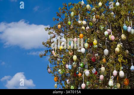 A decorated tree is adorned with colorful Easter eggs, set against a beautiful bright blue sky filled with fluffy white clouds. Stock Photo