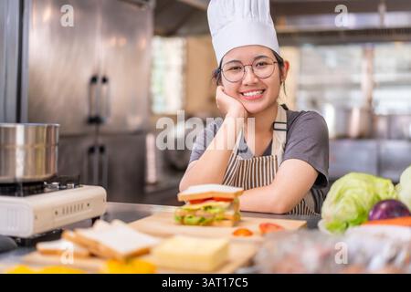 A cheerful young female chef wearing a white hat and striped apron smiles while resting her head on her hand in a commercial kitchen. In front of her Stock Photo
