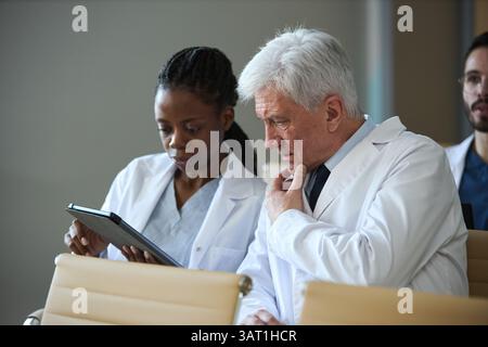 Medical professionals discussing patient records on a digital device during meeting. Interaction between colleagues exchanging information in healthca Stock Photo