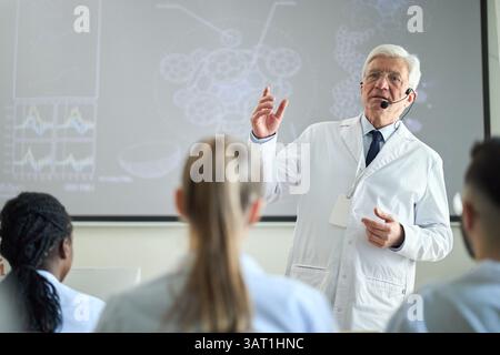 Elderly scientist explaining complex data during seminar while wearing lab coat. Diverse group of professionals attentively listening in classroom, en Stock Photo