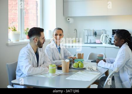 Group of three medical professionals discussing work while seated around table in modern break room. They are sharing ideas over coffee and snacks, fo Stock Photo
