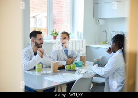 Group of medical professionals enjoying coffee and snacks in a break room while discussing work. Diverse team members in a friendly and relaxed enviro Stock Photo