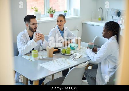 Group of three medical professionals discussing ideas seated at table in hospital cafeteria during break, wearing lab coats and holding coffee mugs Stock Photo