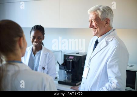 Group of diverse medical professionals in lab coats, smiling and engaging in conversation in a hospital setting. Senior doctor interacting with younge Stock Photo