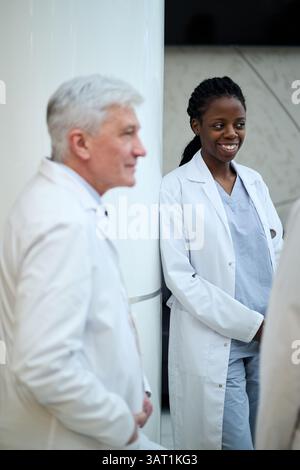 Medical professionals in conversation while standing in hospital. Smiling female doctor wearing white coat and scrubs. Male doctor with gray hair list Stock Photo