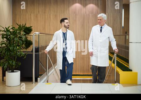 Two doctors having a conversation in a contemporary hospital environment while wearing lab coats. One of them is a senior doctor with white hair, and Stock Photo