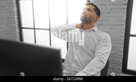 Hispanic man experiencing neck pain while working in an office setting, dressed in a white shirt against a brick wall background, highlighting discomf Stock Photo