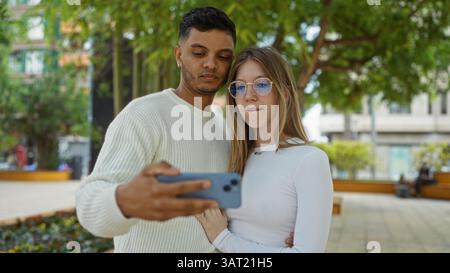 Couple taking selfie in urban park with woman in glasses and man in white sweater surrounded by trees and buildings capturing a moment of togetherness Stock Photo