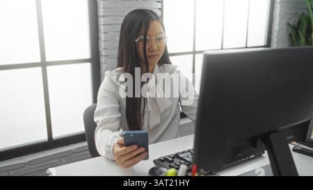 Woman working in a modern office looking focused as she engages with her computer and smartphone, an asian professional combining technology in a prod Stock Photo