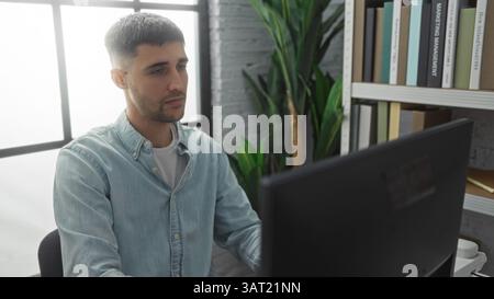 Young hispanic man working at desk indoors in modern office with books and plants, focused on computer screen, displaying an attractive and profession Stock Photo