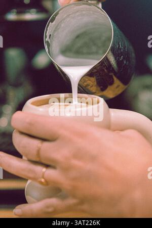 Close-up of hands pouring frothed milk from a stainless steel pitcher into a white cup of espresso, creating latte art Stock Photo