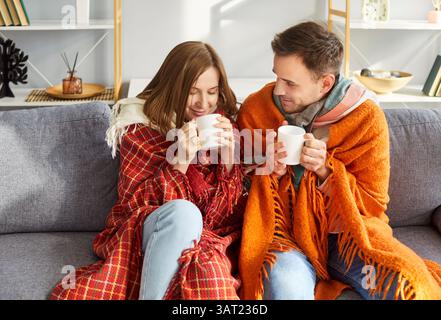 Smiling young couple sitting on sofa under woolen blankets, drinking hot tea and warming up at home. Stock Photo