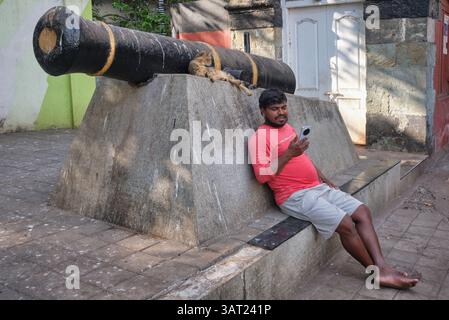 An Indian man sits next to an old cannon marking the entrance to Sassoon Docks in Colaba, Mumbai, India, looking at his mobile phone Stock Photo
