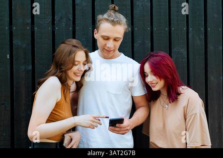 Three friends smiling while looking at a smartphone outside Stock Photo
