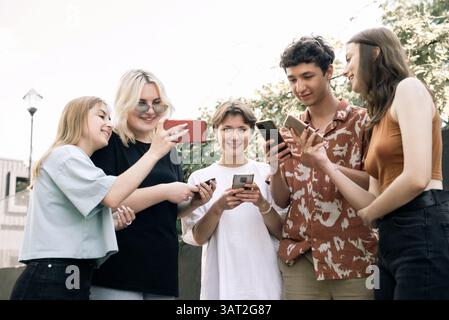 Teen friends smiling while using smartphones outside Stock Photo