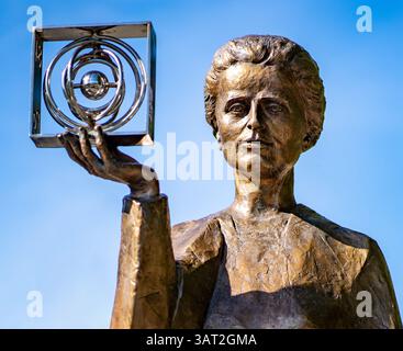 A bronze statue of Marie Curie holding a metallic cube, symbolizing science and innovation, against a clear blue sky. Stock Photo