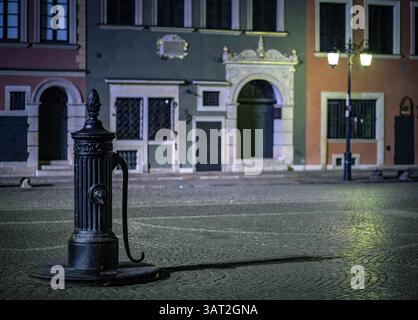 A vintage street pump stands in an empty cobblestone square at night, illuminated by soft streetlights. The surrounding buildings feature classic arch Stock Photo