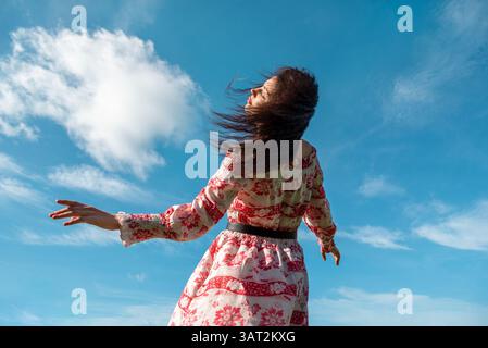 Young woman in a red dress leaning backward toward the camera and enjoying the morning sunlight and wind Stock Photo