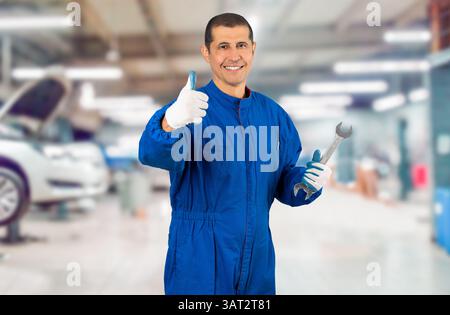 Mechanic posing with a wrench in his hand and gesturing thumbs up at repair shop. Stock Photo