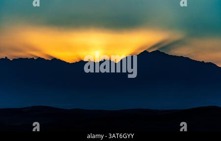 A stunning sunset behind a mountain range, with rays of light breaking through the peaks. The sky transitions from deep blue to warm orange and yellow Stock Photo
