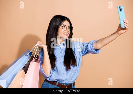 Young woman in casual trendy attire shopping with colorful bags takes a selfie against a beige background, expressing joy and style. Stock Photo