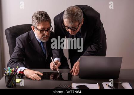 Two senior businessmen in formal suits discussing strategy at office desk using laptop and tablet in modern workplace. Stock Photo