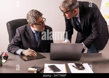 Two senior managers in formal attire working together at office desk with tablet and laptop, analyzing digital documents in a modern business setting. Stock Photo