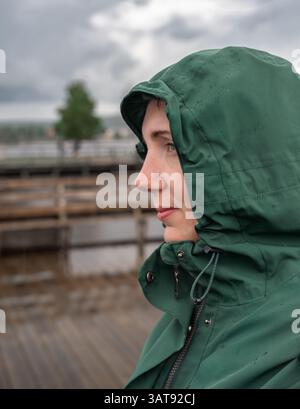 Beautiful woman wearing hooded jacket stands in the rain by a lake. Raindrops cling to her jacket as she gazes thoughtfully at the scenery around her Stock Photo