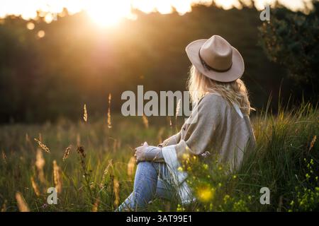 Woman with poncho and hat spending time in nature for mental wellbeing and improve mindfulness. Relaxation on meadow during sunset Stock Photo