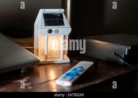 A solar-powered lantern with a visible light bulb, placed on a wooden table next to a laptop and a remote control. The lantern emits a warm glow, crea Stock Photo
