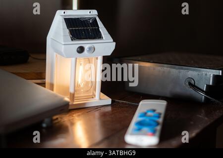 A solar-powered lantern emits a warm glow on a wooden table, surrounded by electronic devices and a remote control, showcasing a blend of modern techn Stock Photo