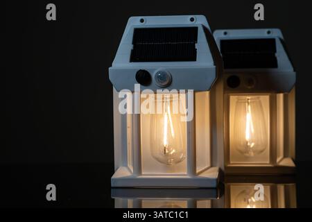 Two solar-powered lanterns with warm glowing bulbs, set against a dark background. The lanterns have a modern design, showcasing their transparent gla Stock Photo
