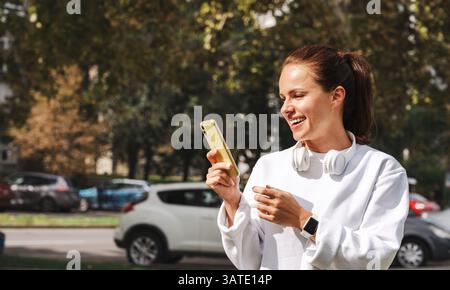 Young woman in white casual sweatshirt using smartphone on city street with headphones around neck Stock Photo
