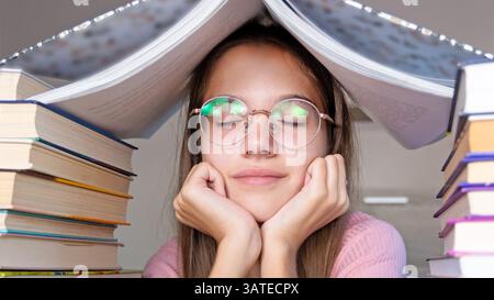 Relaxed Student Resting Amidst Books. A young student wearing glasses sits with his eyes closed under an open book, surrounded by tall stacks of Stock Photo