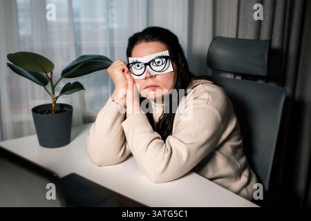 Tired Caucasian woman freelancer feeling stressed from work problems. Irritation burnout. Painted eyes on adhesive notes glued to face. Not having eno Stock Photo