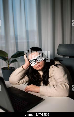 Tired Caucasian woman freelancer feeling stressed from work problems. Irritation burnout. Painted eyes on adhesive notes glued to face. Not having eno Stock Photo