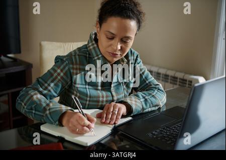 A woman focuses on her tasks, using her laptop and writing notes, seated in a comfortable home workspace. The image reflects a dedicated and productiv Stock Photo
