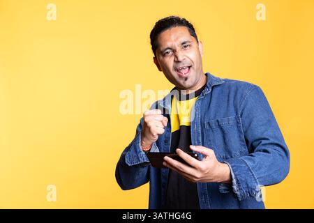 Pleased indian guy cheering after winning game on gaming console, standing against orange background. Delighted young adult bragging after being victorious in video gaming competition. Stock Photo