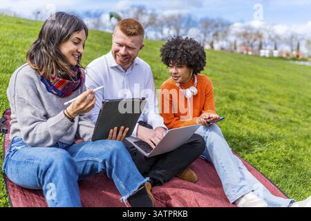 Three university students collaborating on a project, using a digital tablet and laptop while sitting on a blanket in a park Stock Photo