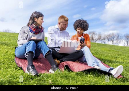 Three students collaborating on a project, using laptop, tablet, and smartphone, enjoying sunny weather in a park Stock Photo