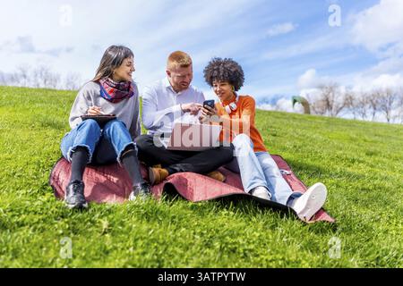 Three students are sitting on grass using laptop and smartphone, enjoying studying together in a park Stock Photo