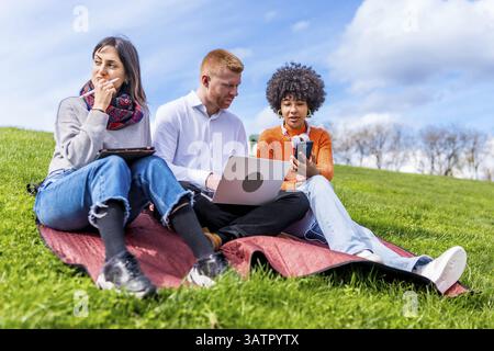 Three students are using laptop, tablet and smartphone, sitting on grass in a park, enjoying spring sunny day Stock Photo
