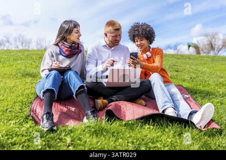 Three university students collaborating on a project outdoors, using a laptop and smartphone while sitting on a blanket in a park Stock Photo