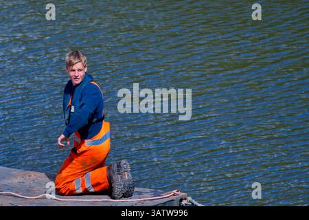 In Amsterdam, a working man in bright orange overalls and blue jacket kneels with resolve on a dock by canal water; his task defines urban maintenance Stock Photo