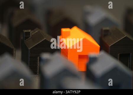 A bright orange miniature house stands out among dark 3D-printed house models, symbolizing real estate choices, market trends, and housing prices. Thi Stock Photo